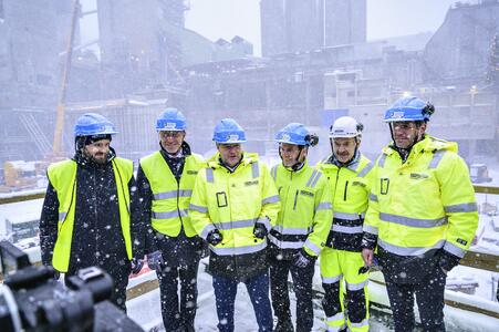 Group of men in work clothes and helmets stand in front of a cement plant