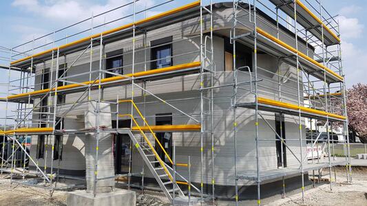 A two-story building under construction, surrounded by scaffolding. The exterior insulation and facade elements are visible, suggesting the building is nearing completion. No workers are present in the scene.