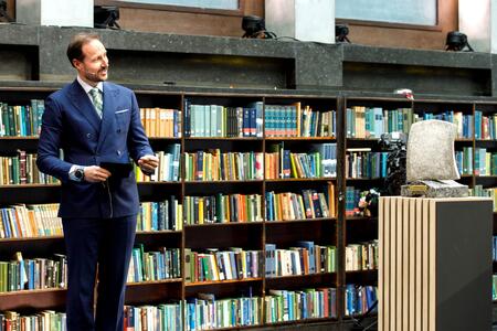 Eine Person in einem blauen Anzug steht vor einem Bücherregal voller bunter Bücher, mit einer abstrakten Skulptur auf einem Podest rechts