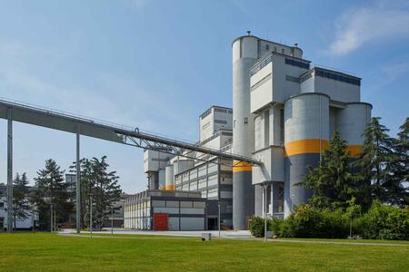 Image of a large industrial cement factory with multiple vertical silos, some with orange bands, under a clear blue sky. A conveyor belt connects two sections at the top. Lush greenery in the foreground.