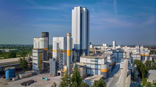 Image of a large cement plant with multiple buildings of varying heights. The most prominent structure is a tall, white building with vertical blue stripes. Several large cylindrical silos and rectangular buildings with visible piping and ductwork are also present.