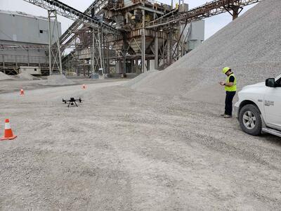 Eine Drohne steht auf einer Kiesfläche in einem Steinbruch. Im Hintergrund befinden sich große Kieshaufen, Industrieanlagen und Förderbänder. Rechts steht eine Person in Schutzkleidung, die die Drohne steuert.