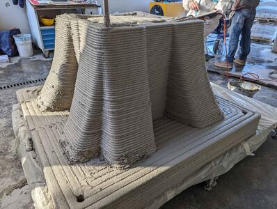 Ein 3D-Drucker erstellt eine großformatige Struktur in Form einer umgekehrten Pyramide aus Beton und zeigt die geschichtete Drucktechnologie.
