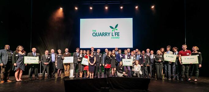 Award recipients lined up on stage at ‘The Quarry Life Award’ ceremony holding checks with prize amounts, with event logo displayed on screen behind.