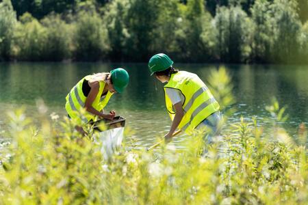 Zwei Frauen mit Helm und Warnweste stehen mit Keschern in einem See, im Vordergrund Uferbegrünung