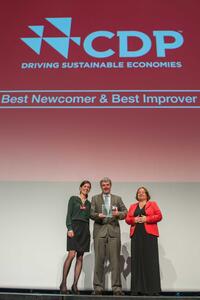 Two women and a man are standing on a stage. The man is holding an award in his hands. On the wall behind it says ‘CDP, best newcomer & best improver’.