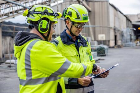 Zwei Mitarbeiter in Schutzkleidung stehen in einem Werk und schauen sich gemeinsam Unterlagen an