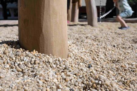 Close-up view of a wooden post planted into the ground, surrounded by light-coloured gravel. In the background, there is a blurred figure of a person engaged in an activity involving a rope or cord.