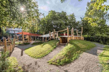Playground featuring wooden structures, grassy mounds, and gravel pathways, surrounded by trees and greenery.