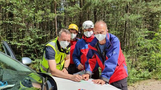 Four men wearing FFP2 masks lean against the bonnet of a car and look at a map, with a forest in the background.