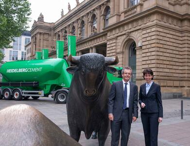 Mann und Frau in Businesskleidung neben einer Stier-Skulptur vor der Börse Frankfurt, im Hintergrund ein grüner Silozug
