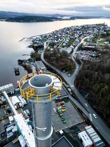 An aerial view of a coastal town with a large industrial chimney in the foreground. The chimney has a yellow safety railing at its top.
