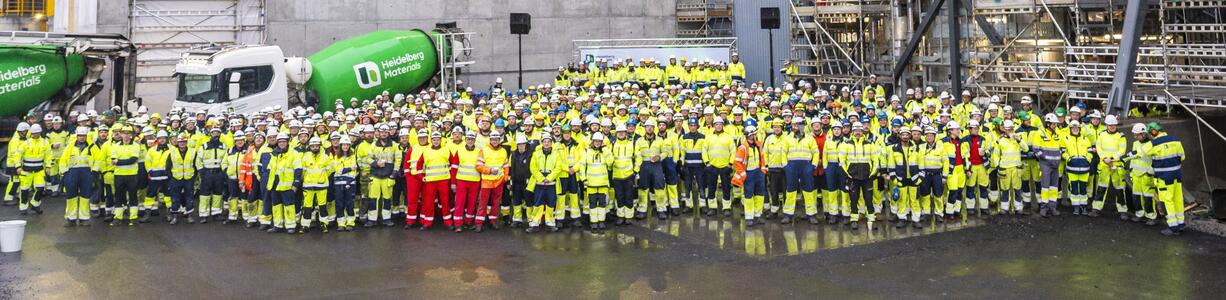 Group picture with a lot of people in neon yellow protective clothing, in the background green and white ready-mixed concrete trucks and industrial buildings