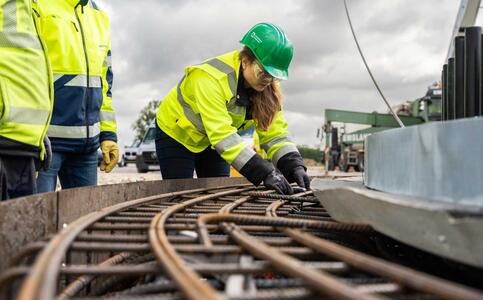 A woman wearing a green hard helmet, protective goggles and protective clothing is working on a reinforcement bar inside a formwork. Two other people in protective clothing are standing next to her; heavy construction equipment can be seen in the background.