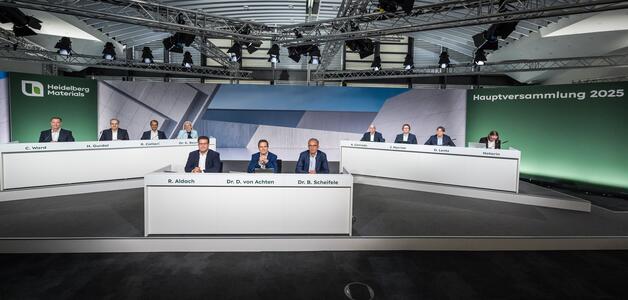 Podium mit elf Personen in formeller Kleidung, die hinter weißen Tischen sitzen; zentrale Figur (Frau) spricht in ein Mikrofon. Im Hintergrund Logo und Schriftzug „Hauptversammlung 2025“ auf grüner Fläche; deckenmontierte Beleuchtung und Kameras sichtbar.