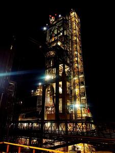 A brightly illuminated industrial structure at night, featuring a tall vertical tower with interconnected platforms, machinery, and metallic frameworks. Lights highlight key areas, and the dark sky contrasts with the glowing structure, emphasizing its intricate design and height.