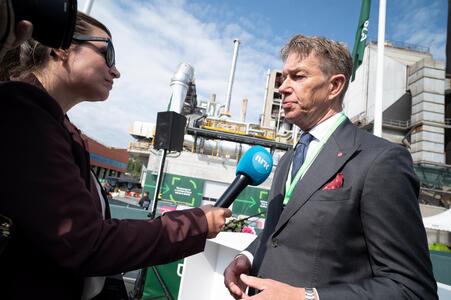 A reporter holds a microphone bearing a news logo while interviewing Terje Aasland in a suit and green lanyard. The background features a cement facility with visible structures, pipes, and a partially cloudy sky.