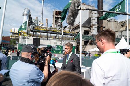 Terje Aasland in a suit and green tie speaks to a group of journalists with cameras and microphones in an industrial setting. The background includes large pipes, machinery, and green flags, indicating a corporate event at a cement facility.