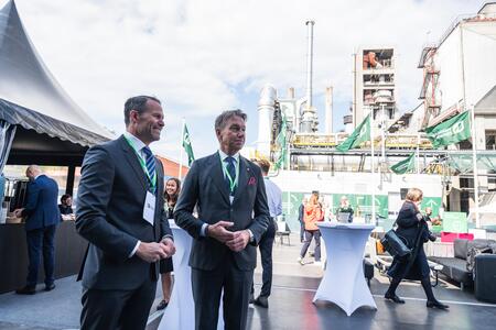Two men in formal business attire, wearing lanyards, stand conversing at the Brevik launch event. Behind them, a cement facility with metal structures and pipes is visible. White cocktail tables are scattered around, with a few other attendees mingling in the background. The sky is partly cloudy, and a large event tent is seen on the left.