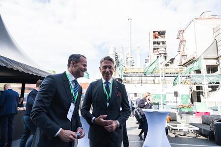Two men in business suits with green lanyards smile and converse outdoors at the Brevik launch event. In the background, industrial equipment and structures are visible under a partly cloudy sky, along with event tables and attendees.