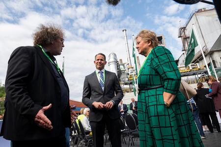 Three individuals are standing outdoors at an industrial facility under a partly cloudy sky. On the left, a man with curly hair gestures while speaking. In the center, a man in a suit and glasses smiles, holding his hands together. On the right, a woman in a green plaid coat listens attentively. Behind them, industrial structures with metallic pipes and tanks are visible, suggesting a discussion related to the facility.