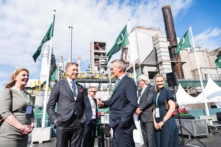 Group of well-dressed individuals standing outdoors at an industrial site with chimneys and equipment in the background. Green flags with the Heidelberg Materials logo are visible, and the individuals are smiling and engaged in conversation under a bright sky.