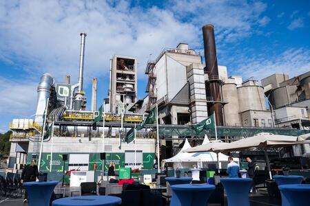 Cement plant with multiple large chimneys, pipes, and steel structures under a partly cloudy blue sky. In the foreground, outdoor event setup with tables, chairs, and white canopy tents is visible. People are scattered around the area, indicating an event.