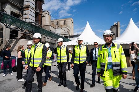 Group of people, including H.R.H. Crown Prince Haakon of Norway, wearing yellow high-visibility vests and white helmets walking at the Brevik cement plant with large white tents in the background, under a clear blue sky.