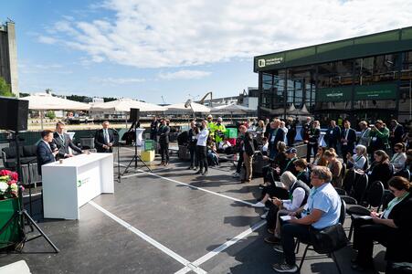 The Brevik launch event with Terje Aasland at a white podium on the left addressing a seated audience on the right. Attendees include media and onlookers, with cameras and microphones visible. The background features a modern green building, umbrellas, and industrial structures under a partly cloudy sky.