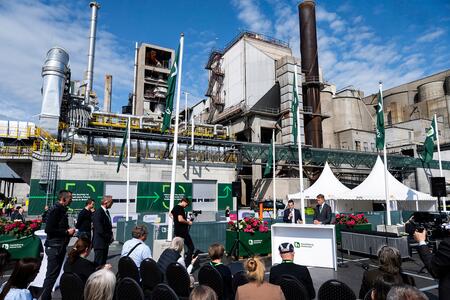 People seated outdoors at a cement facility during the Brevik launch event, with speakers at a podium in the foreground and plant structures, chimneys, and pipes in the background. Green and white banners and flower arrangements decorate the area under a partly cloudy sky.