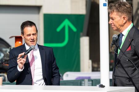 Dominik von Achten and Terje Aasland in suits are at the Brevik Launch event, one speaking into a handheld microphone. They are positioned at a white podium or table, outdoors, with a green upward arrow sign in the background. A microphone stand is visible on the right side.