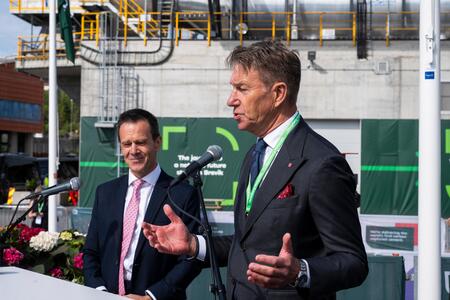 Von Achten and Aasland in formal attire stand behind microphones at the Brevik launch event, with industrial equipment, green construction banners, and flowers visible in the background. The person on the right is gesturing with their hands while speaking.