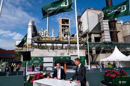 Von Achten and Aasland in formal suits are speaking at the Brevik launch event in front of an industrial plant with large pipes, towers, and equipment under a clear blue sky. Green flags with circular arrows and a logo are displayed prominently. Pink flowers adorn the front of the stage area, and a white canopy is visible on the right. The setting suggests an environmentally focused or corporate event.