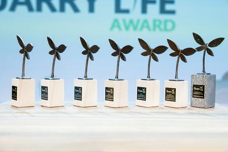 Sieben Trophäen mit stilisierten Blättern auf weißen Sockeln, eine davon mit silbernem Sockel, in einer Reihe auf einem hellen Tisch; im Hintergrund der Schriftzug „LIFE AWARD“.
