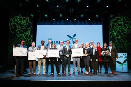 A group of diverse individuals stands on a stage holding large checks and awards, signifying a recognition event. The backdrop features a central white screen displaying a logo with green and blue leaves, accompanied by decorative green line art on both sides. The participants are dressed formally, smiling, with stage lighting illuminating them.