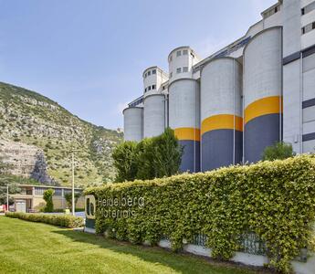 Industrial silos with yellow and gray stripes stand near a hillside, surrounded by greenery on a Heidelberg Materials site.