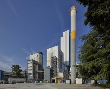 A cement plant with tall silos and a chimney, surrounded by trees and clear skies.