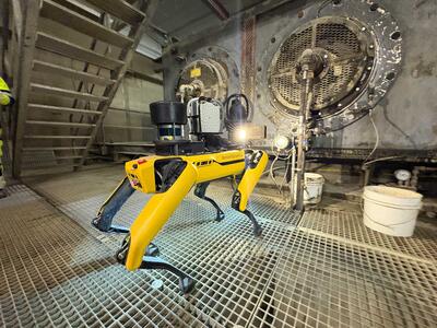 A robotic dog equipped with sensors is inspecting an industrial site with large machinery in the background.