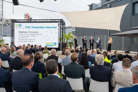 An outdoor corporate event with an audience listening to a panel discussion on stage in front of a presentation screen.