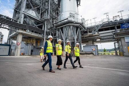 People in safety gear walk in front of an industrial facility.