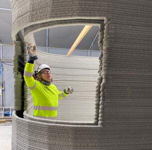 Man in protective clothing and a construction helmet stands in the window of a 3D-printed building