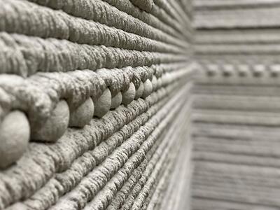 Close-up view of a 3D-printed concrete wall, showing the visible layers of the printed material. The foreground is sharply focused, gradually blurring towards the background, creating a sense of depth.