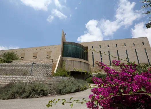 A modern building with a curved glass facade and a concrete wing-like structure, set amidst greenery and purple flowers under a partly cloudy sky