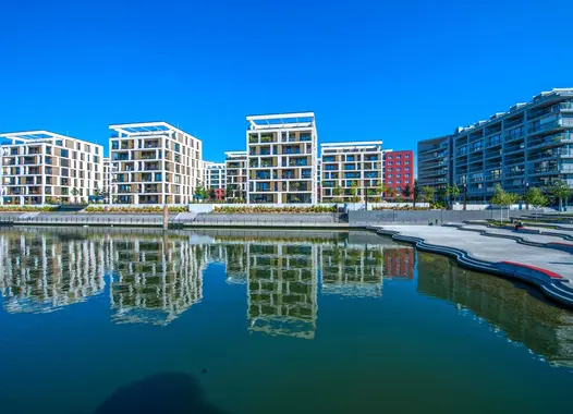 A contemporary urban riverside development with several white structures adorned with balconies and red highlights, mirrored in the tranquil waters of a neighboring river or canal beneath a clear blue sky.
