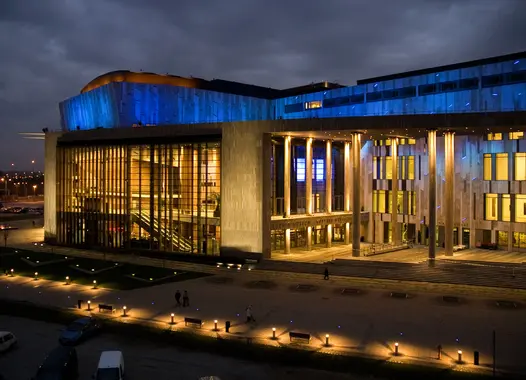 A contemporary building stands out at dusk, its curved roof adorned with blue lights, while warm lights from within reveal its modern design and welcoming atmosphere.