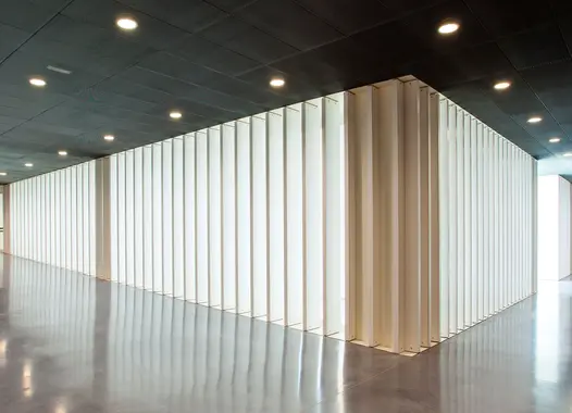 A modern interior featuring a large, rectangular structure made of vertical white panels that allow light to filter through, creating a pattern on the glossy floor, under a dark ceiling with recessed lighting.