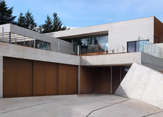 A modern residence with geometric design, combining concrete and wood, featuring large windows and a flat roof, under a partly cloudy sky