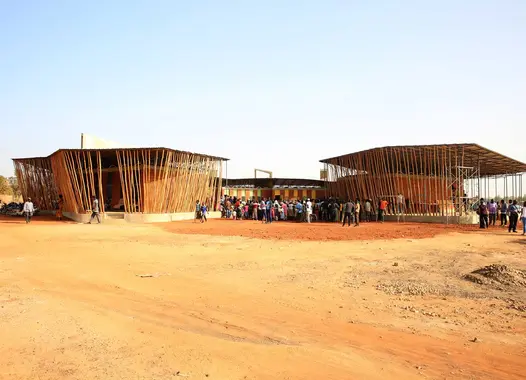 group of individuals standing in front of two large wooden and bamboo structures that are either under construction or designed in an open framework style, with a natural landscape in the background