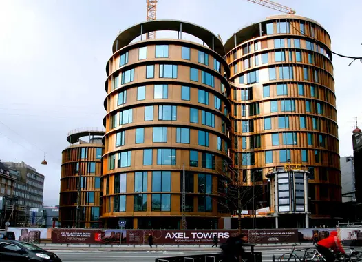 A modern building under construction with a distinctive architectural design featuring curved shapes and a facade with gold and brown hues. The building has multiple floors, large windows, and two visible cranes on top, indicating ongoing work.