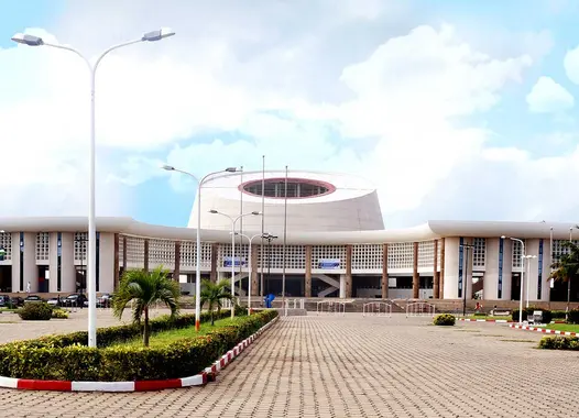 Modern symmetrical building with curved facade and circular windows, flanked by wing-like extensions under an overcast sky, fronted by a geometric plaza.